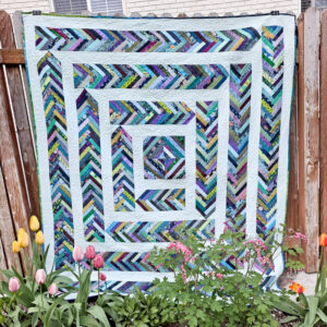 Braid quilt in blues, greens, and purples hanging on a fence with flowers growing underneath