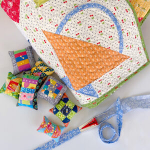 A quilted basket block with a pile of patchwork pincushions and a bias tape maker in use
